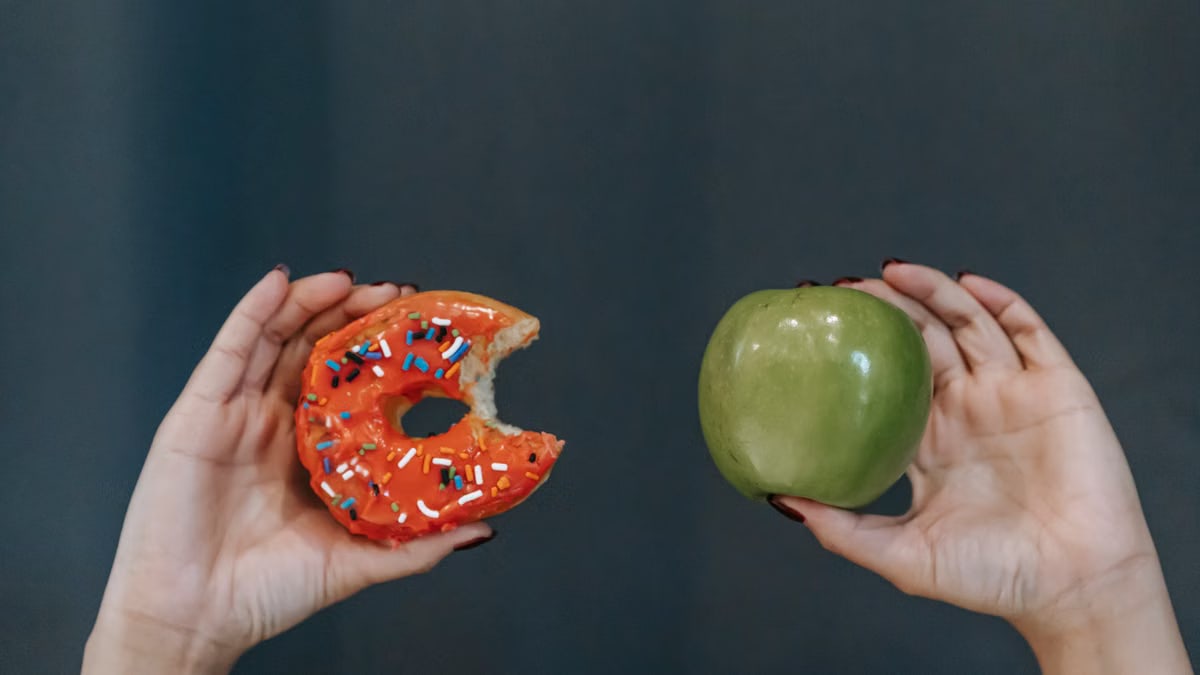 Crop unrecognizable young female demonstrating bitten high calorie doughnut and healthy green apple against gray background